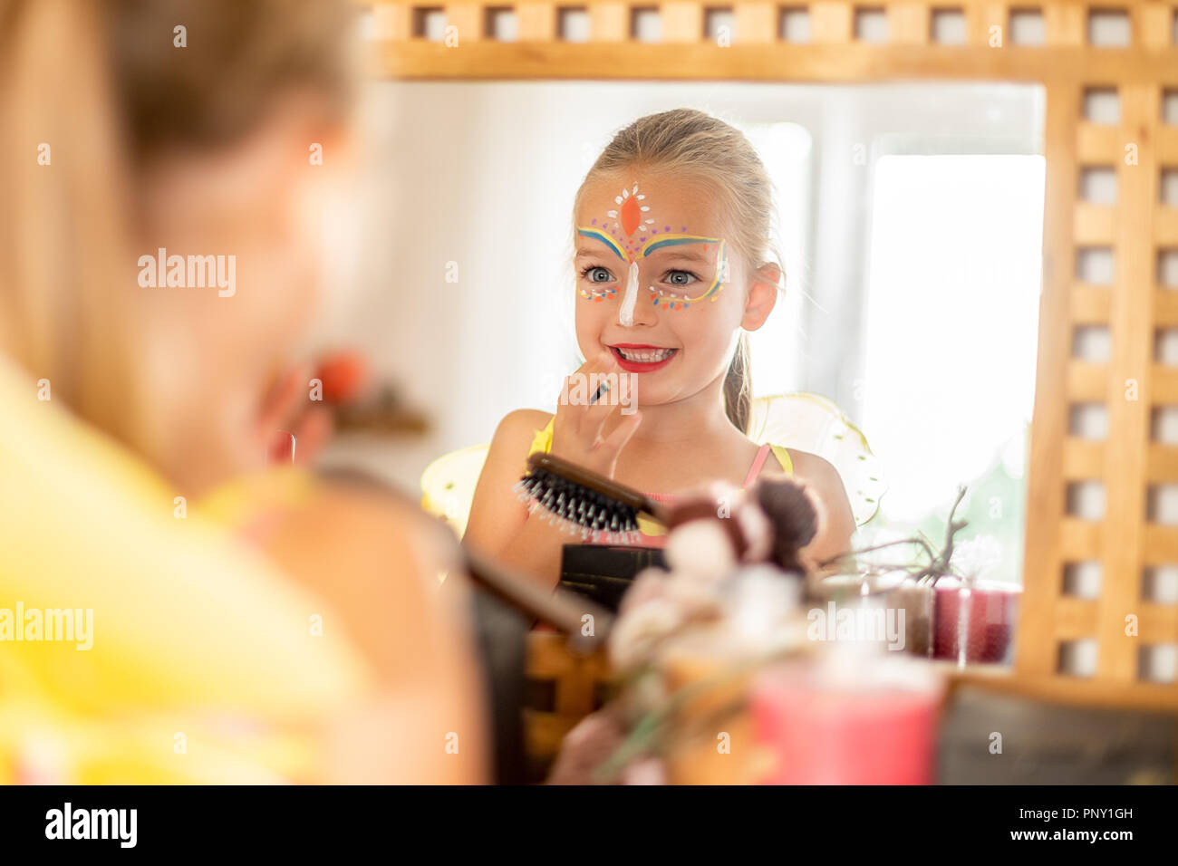 Happy child with face paint and wearing halloween costume looking in the mirror, smiling. Excited young girl looking at herself in the mirror. Stock Photo