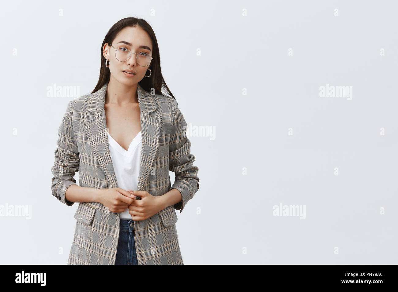 Girl hesitating, wanting tell something important. Portrait of serious unsure female entrepreneur in stylish jacket and glasses, touching fingers and gazing at camera with doubt over grey wall Stock Photo