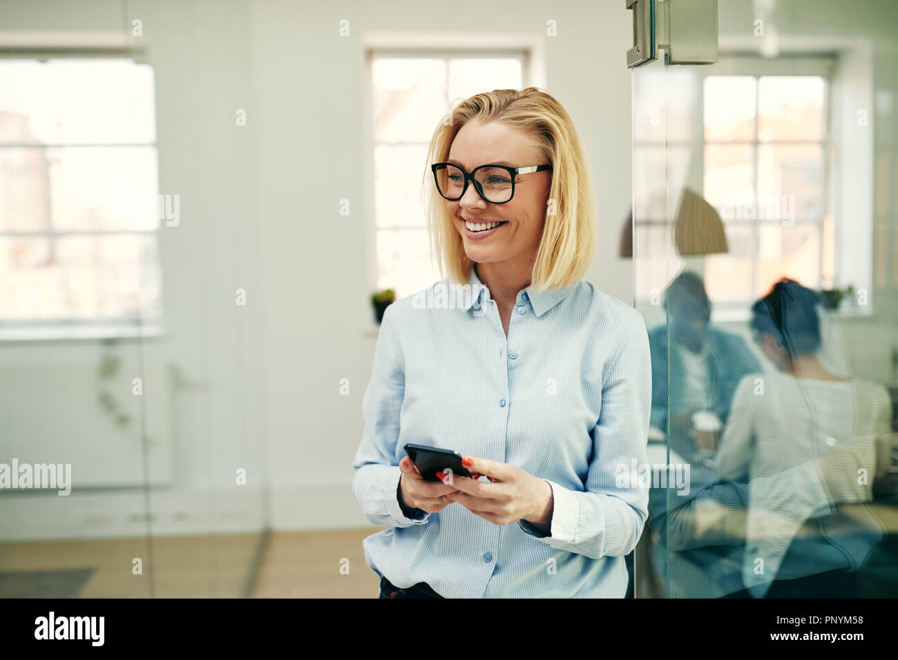 Young businesswoman standing in a modern office and laughing while reading a funny text message on her cellphone Stock Photo