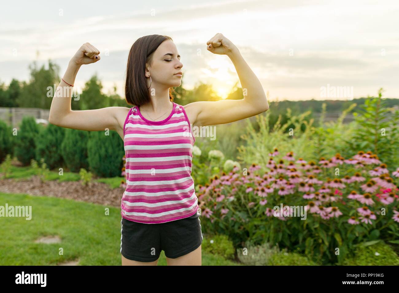 People, power, stamina, strength, health, sport, fitness concept. Outdoor portrait smiling teenage girl flexing her muscles, background green lawn sun Stock Photo