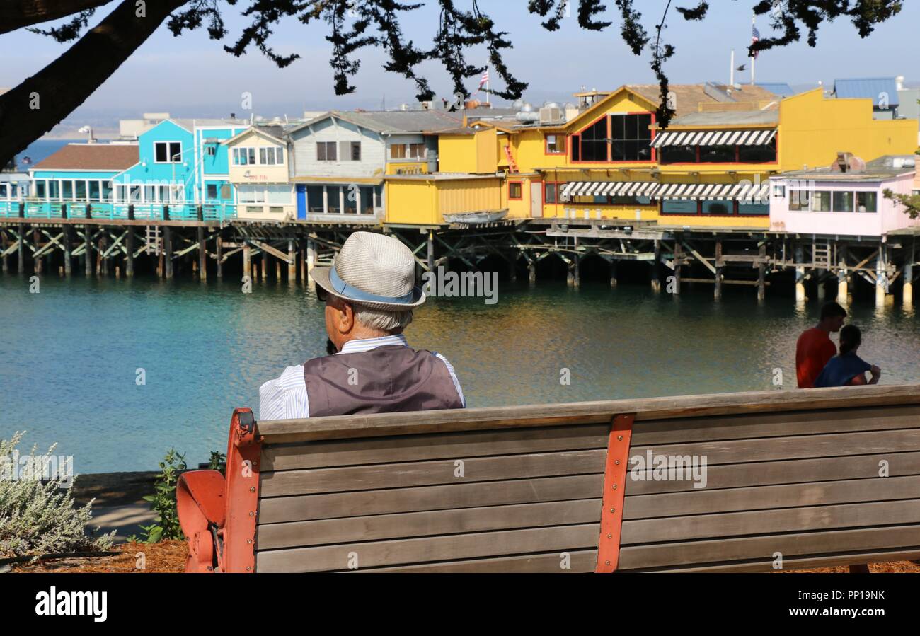An elderly man wearing a dapper hat sits on a bench enjoying the view of Fisherman's Wharf, Monterey, CA. Stock Photo
