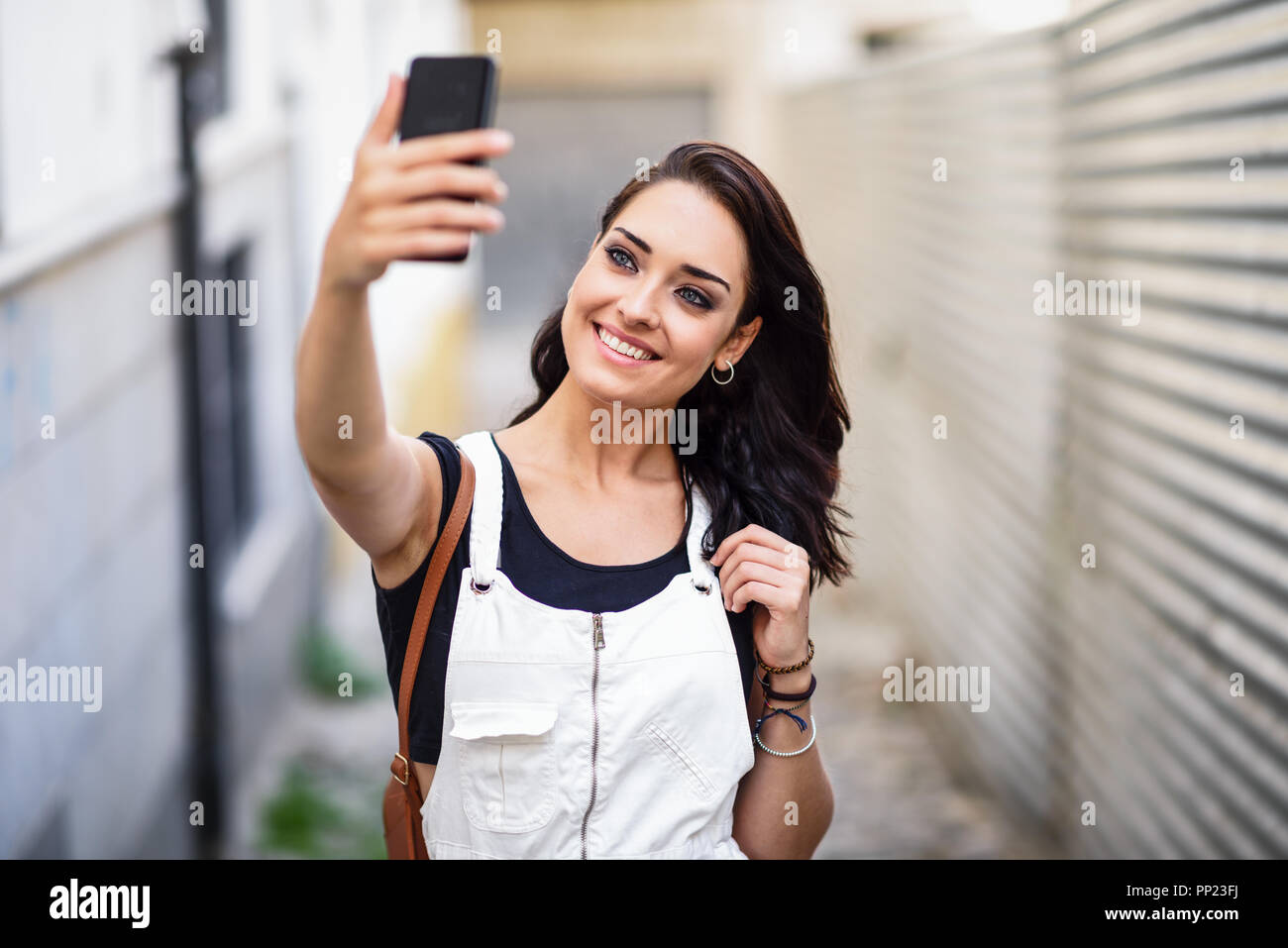 Young woman taking selfie photograph with her smart phone outdoors. Girl wearing denim dress in urban background. Technology concept. Stock Photo