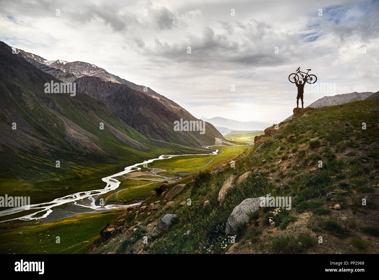 Epic shot of Man holding his mountain bike on the hill in silhouette with the river in the mountain valley background. Stock Photo