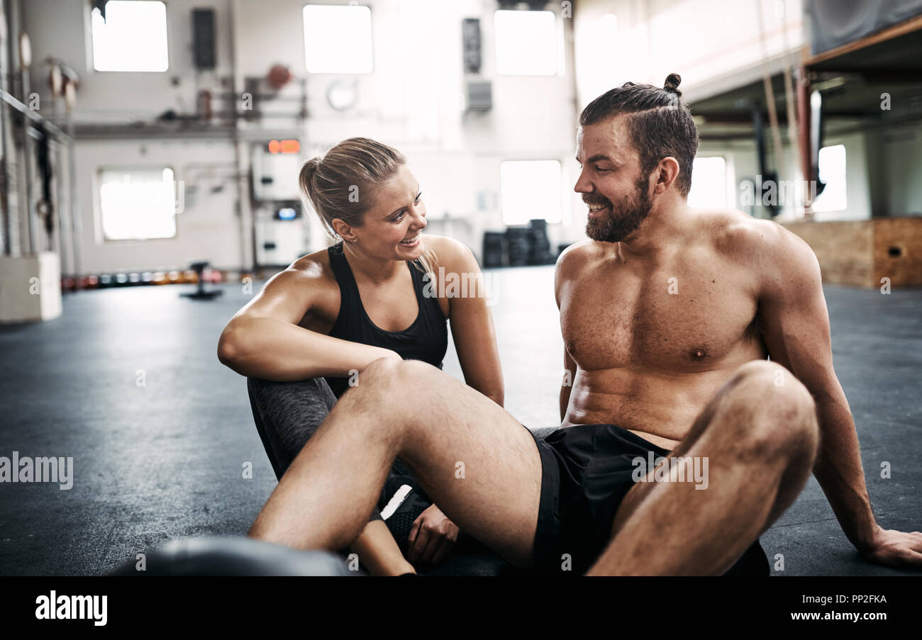 Fit young couple in sportswear sitting together on a gym floor smiling and talking while taking a break from working out Stock Photo