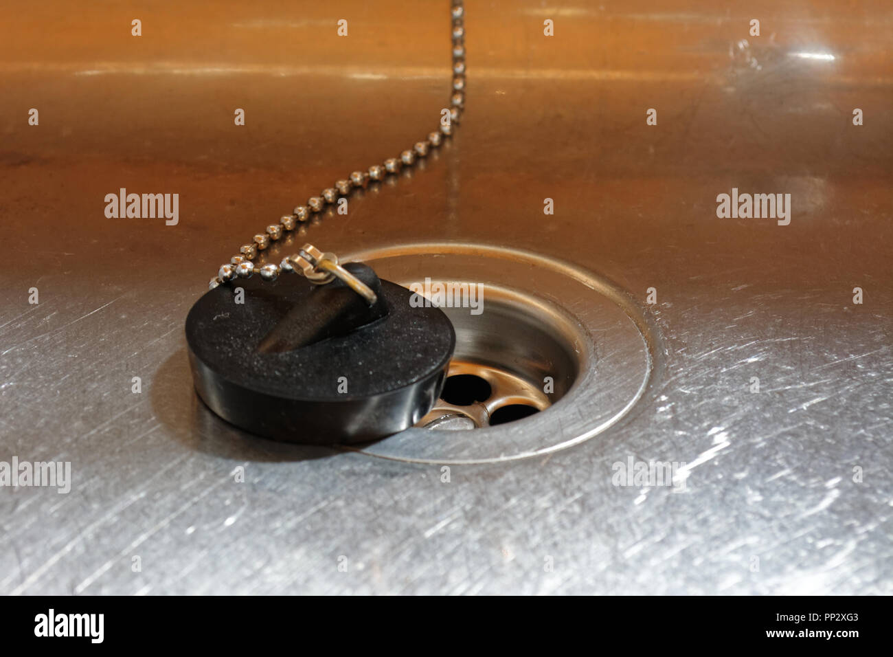 Black plug on beaded chain sits next to a plug hole in a stainless steel kitchen sink. Stock Photo