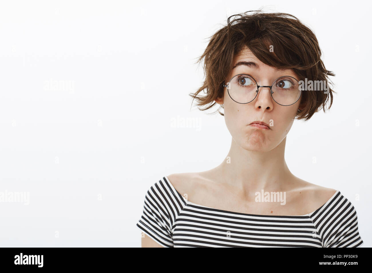 Wow, it is amazing but unexpected. Impressed cute and funny girl in round  glasses with stylish short haircut, pulling face in not bad emotion,  looking left curiously, standing pleased over grey wall