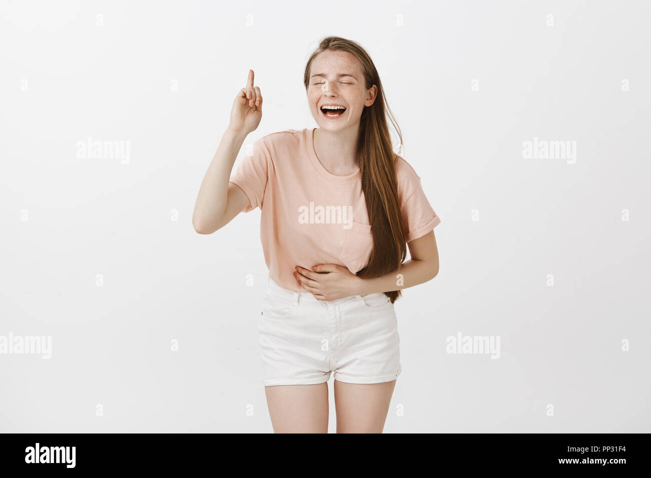 Pretty carefree girl having fun, laughing out loud seeing amusing comedy show, bending from laugh and touching belly, smiling broadly with closed eyes, pointing up with index finger over gray wall Stock Photo