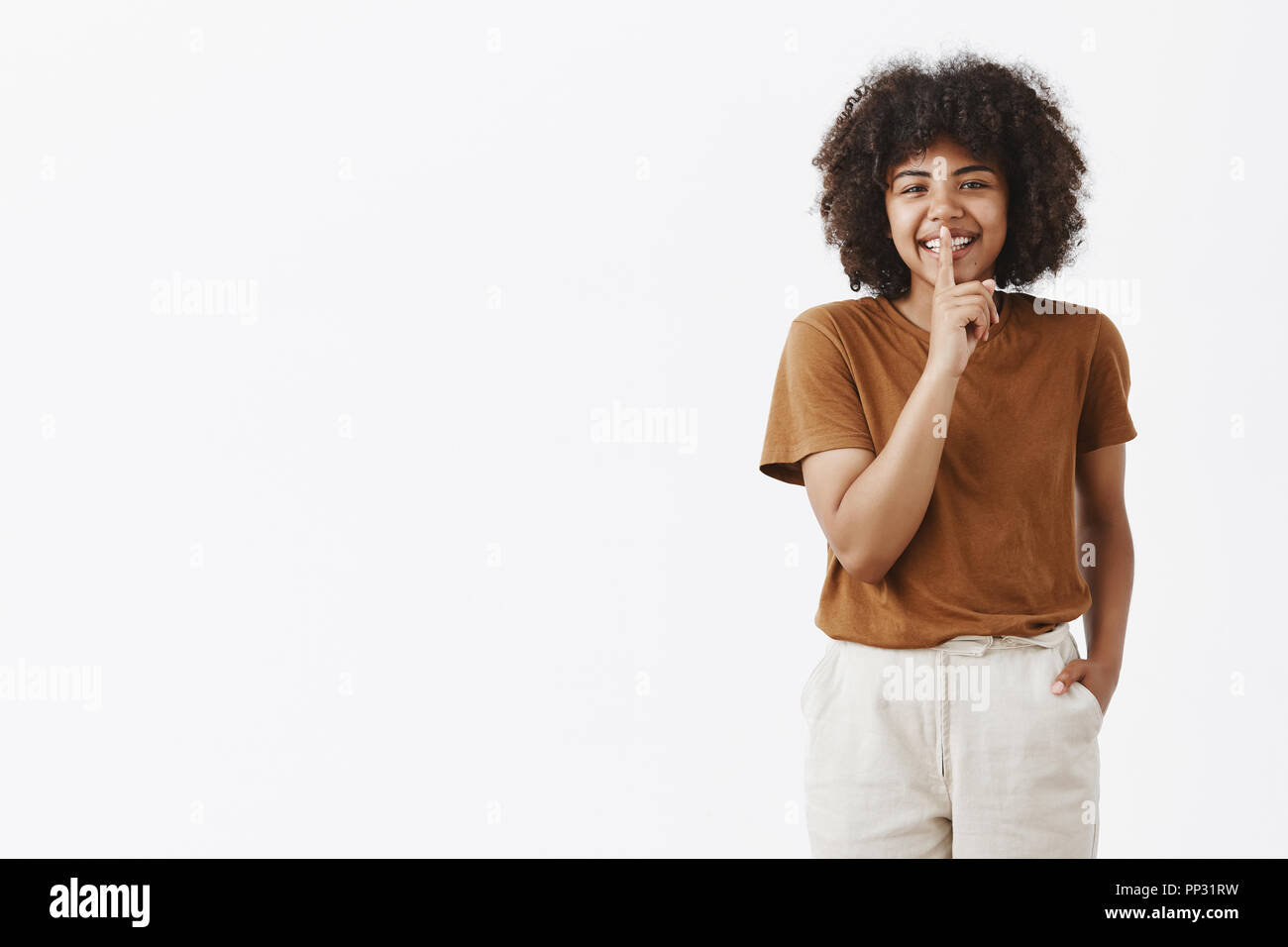 Happy carefree african-american cute teenage girl with afro hairstyle in stylish brown t-shirt and white pants holding hand in pocket casually smiling, showing shush sign with index finger over mouth Stock Photo