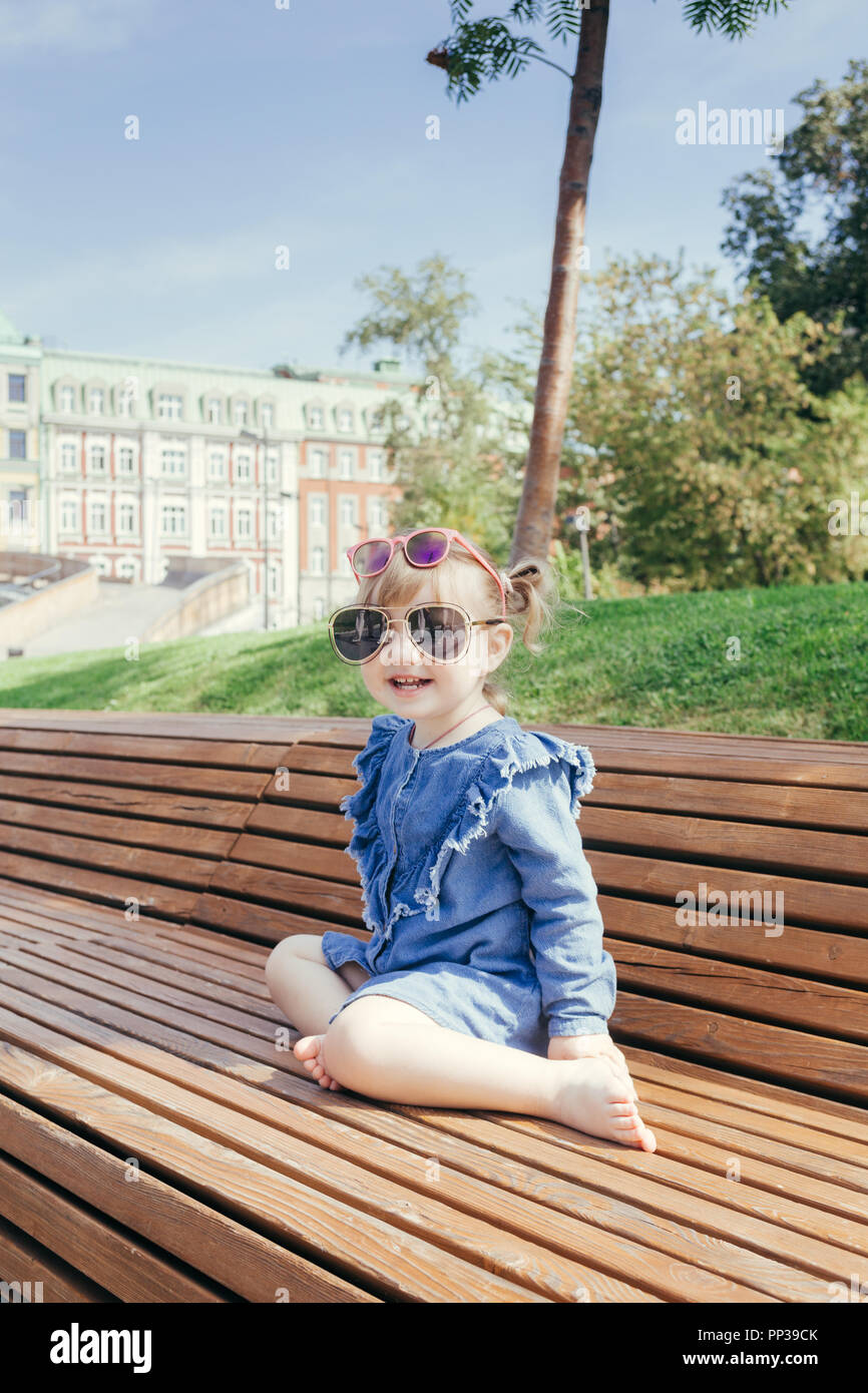 Little girl in sunglasses sitting on the bench in a city park on a warm ...