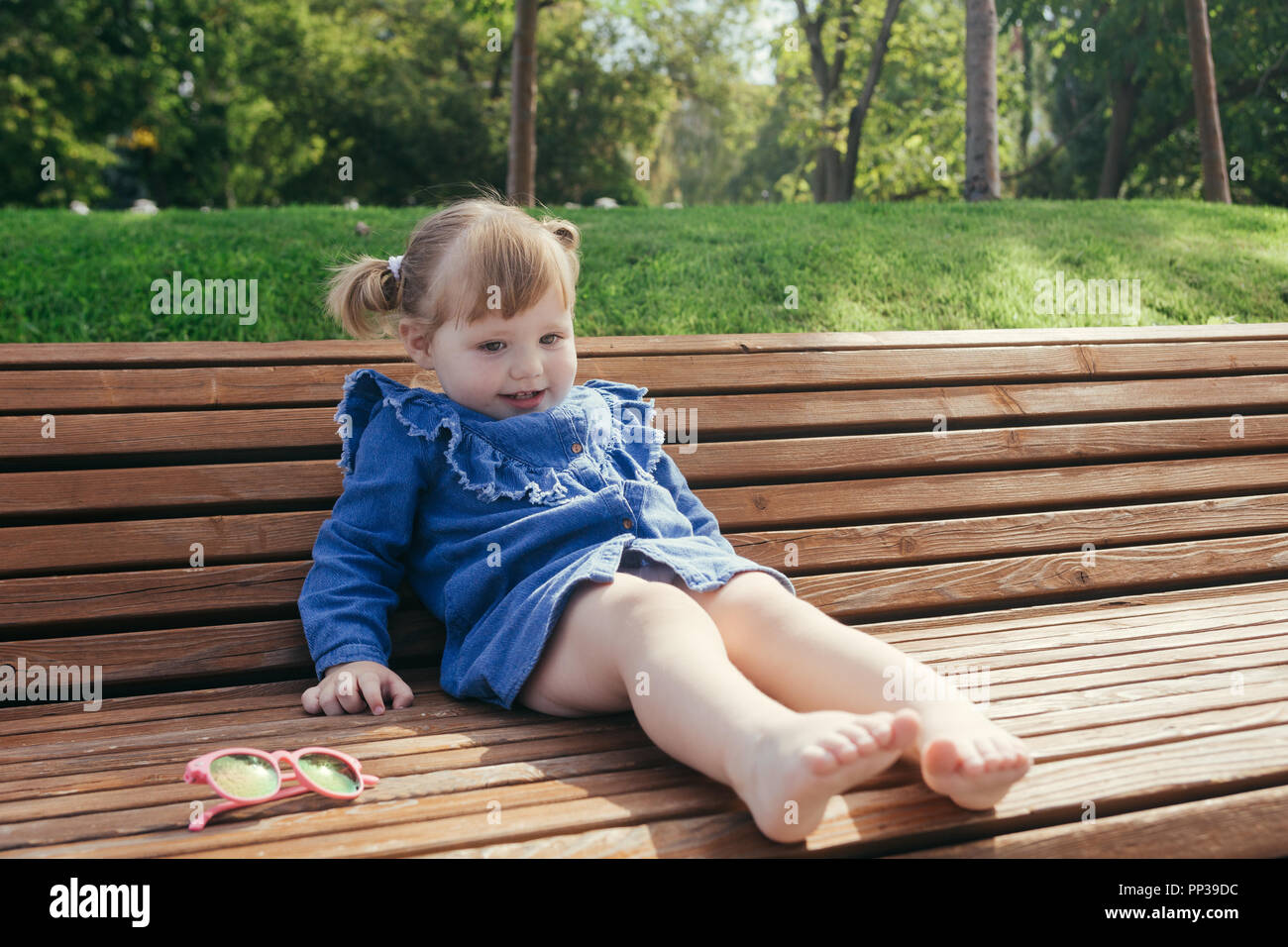 Little girl sitting on the bench in a city park on a warm sunny day ...