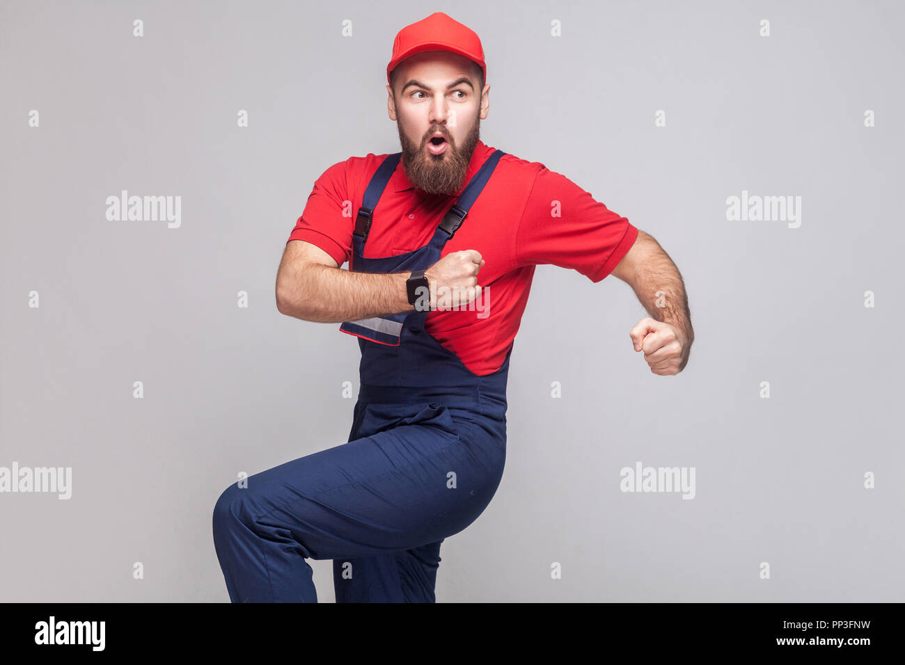 Hurry up! Young amazed handyman with beard in blue overall, red t-shirt and cap are late and starting to run for help on time. Grey background, indoor Stock Photo
