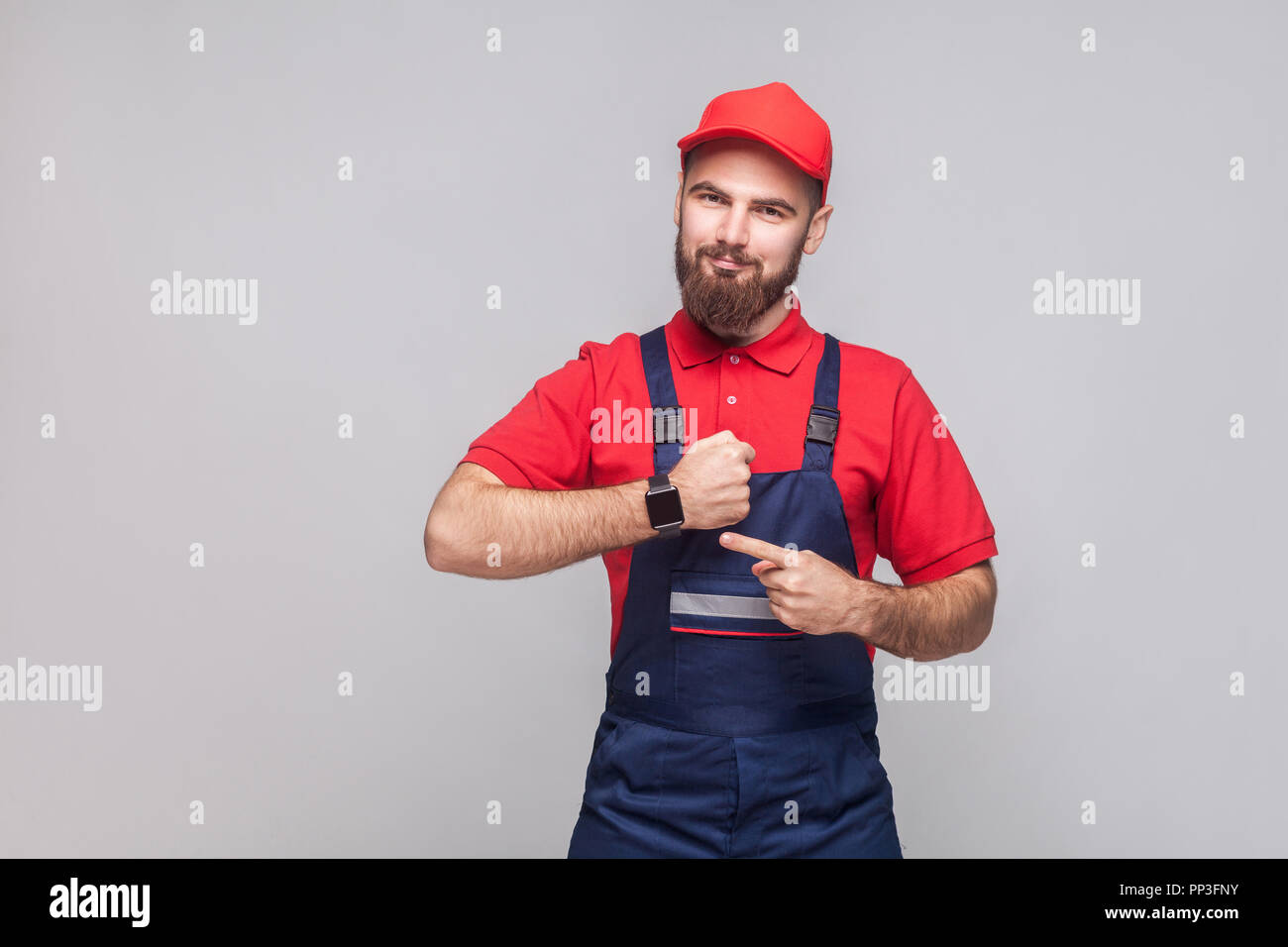We do ontime. Young confident handyman with beard in blue overall and red t-shirt standing and showing time on his wrist watch with smile. Grey backgr Stock Photo