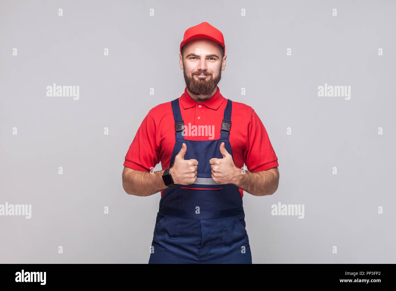Work are done! Portrait of young satisfied cheerful repairman with beard in blue overall, red t-shirt and cap, standing and showing thumps up with smi Stock Photo