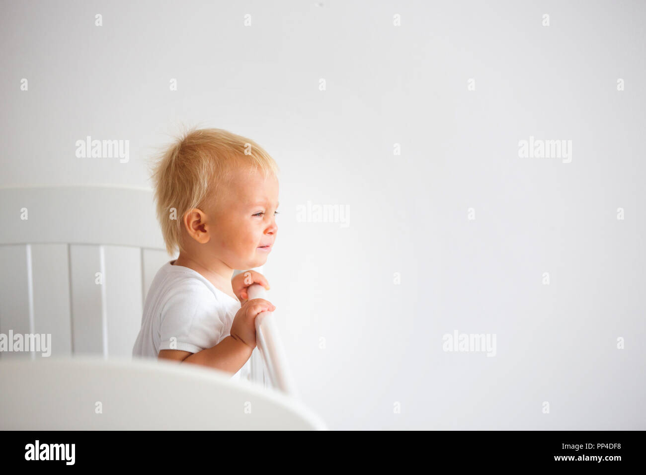 Portrait of bored baby standing in crib. Baby boy stand alone in crib. Sad little baby. Lonely