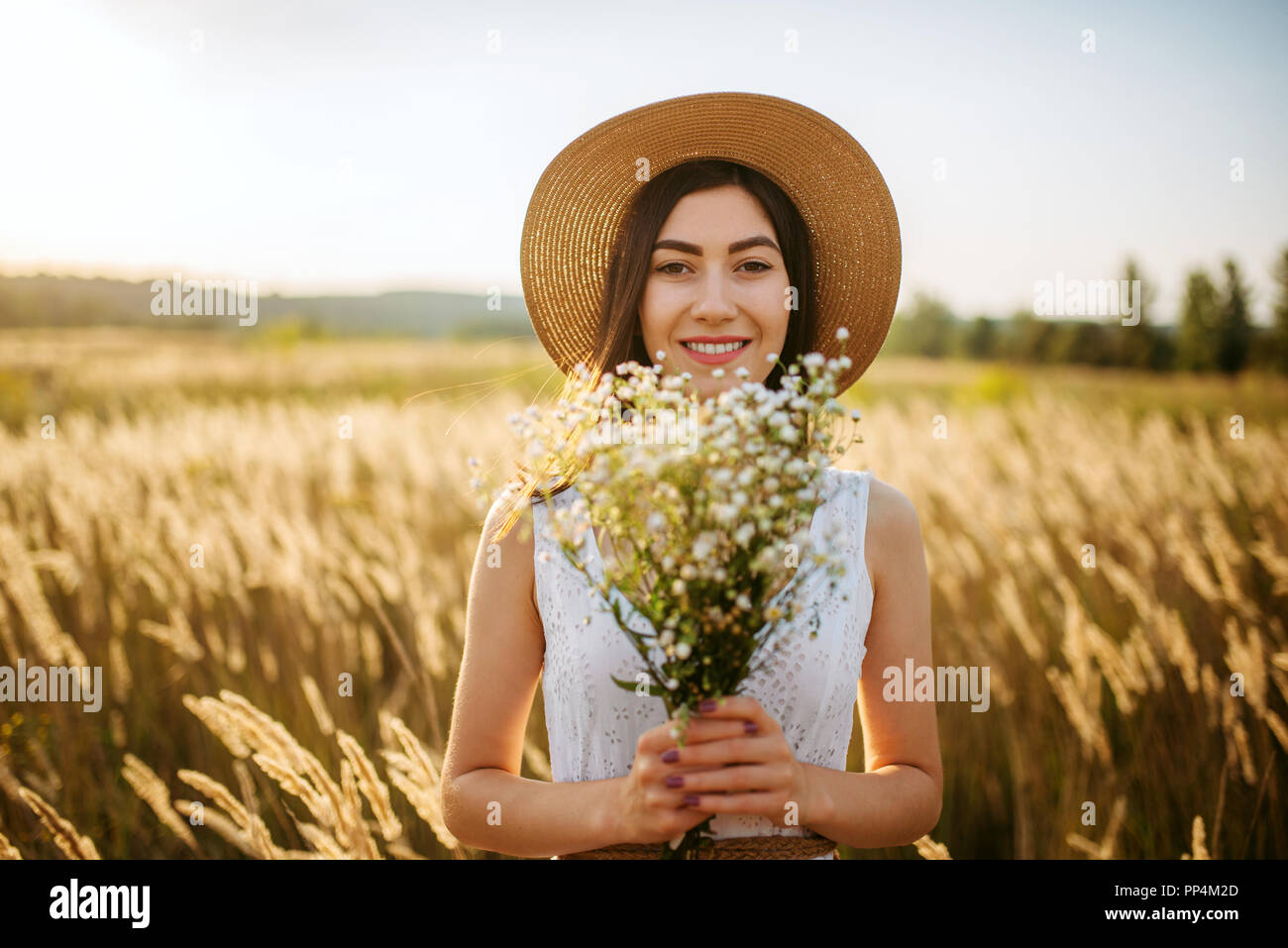 Young woman in white dress and straw hat with a bouquet of wild flowers ...
