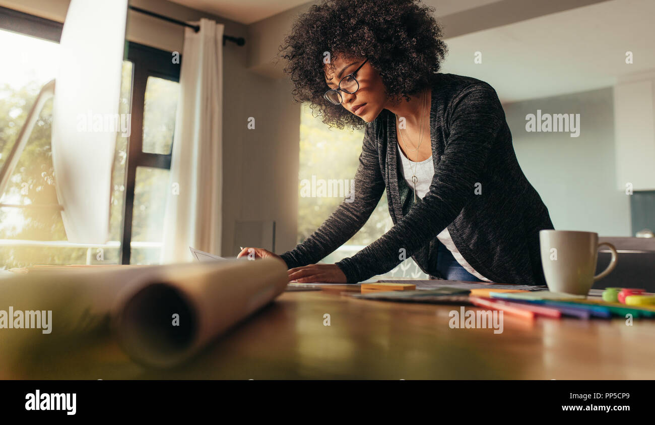 Young female architect working on a project from home office. African woman standing at the table and looking at some documents. Stock Photo