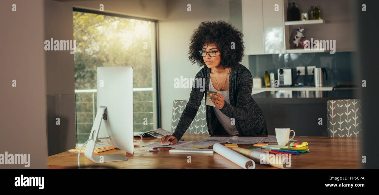 Woman interior designer with color swatch in hand working on computer in the home office. Young african woman standing at the table and looking for a  Stock Photo