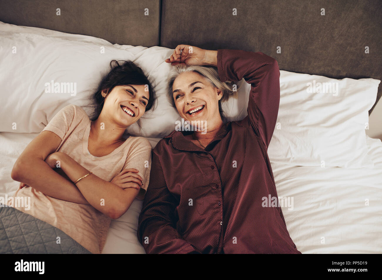 Smiling woman sleeping beside her old mother on bed with arms crossed