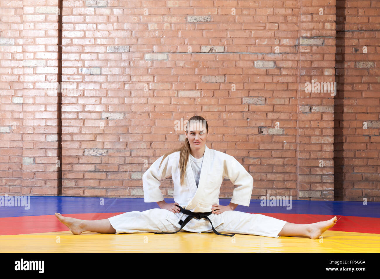 Beautiful athletic karate woman in white kimono with black belt stretching in twine and looking at camera. Japanese martial arts concept. Indoor, stud Stock Photo