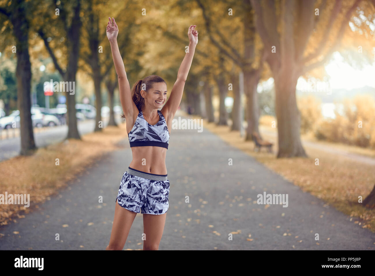 Fit happy slender healthy young woman in sportswear standing in a tree lined avenue raising her arms with a beaming smile Stock Photo