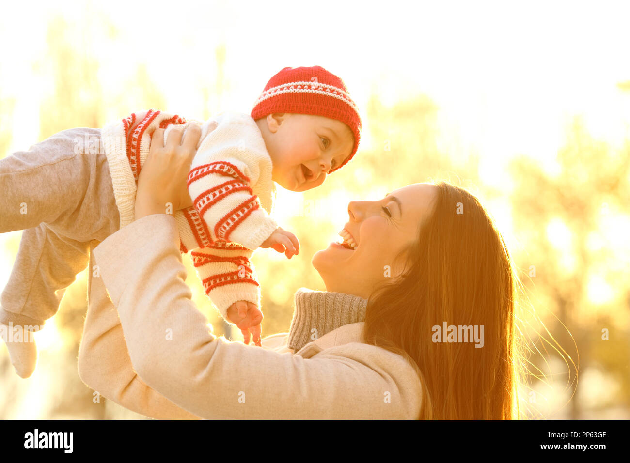 Backlighting portrait of a joyful mother raising her baby outdoors at sunset in winter Stock Photo