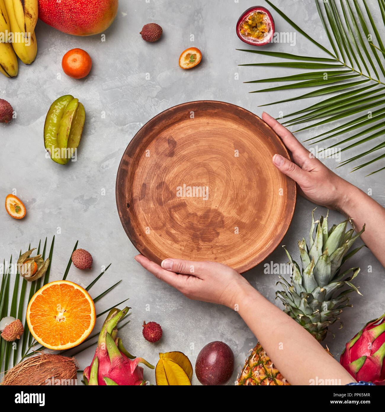 Hands of a woman holding a brown empty plate around a different tropical fruit ripe on a gray concrete background with copy space. Flat lay Stock Photo