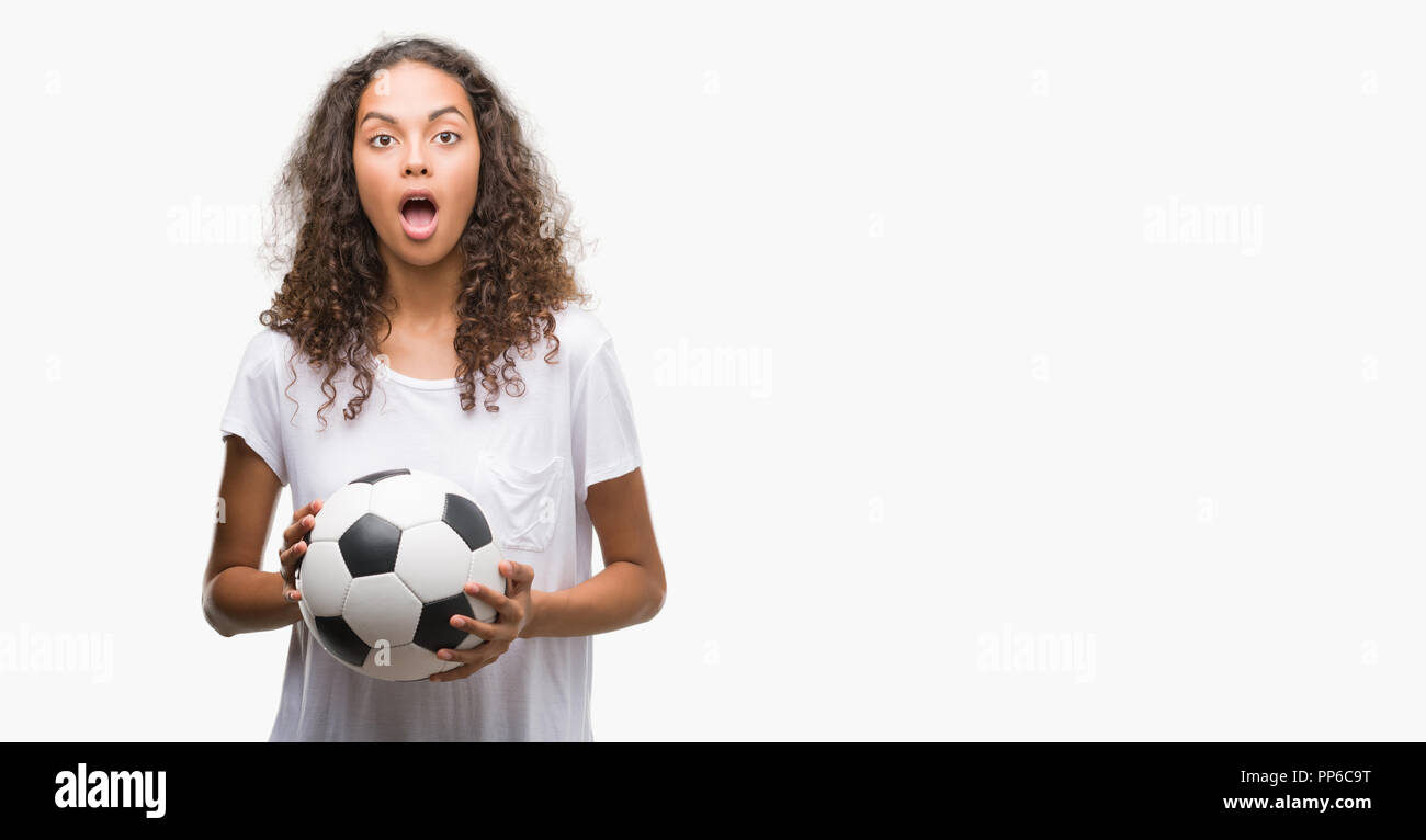 Young hispanic woman holding soccer football ball scared in shock with a surprise face, afraid and excited with fear expression Stock Photo