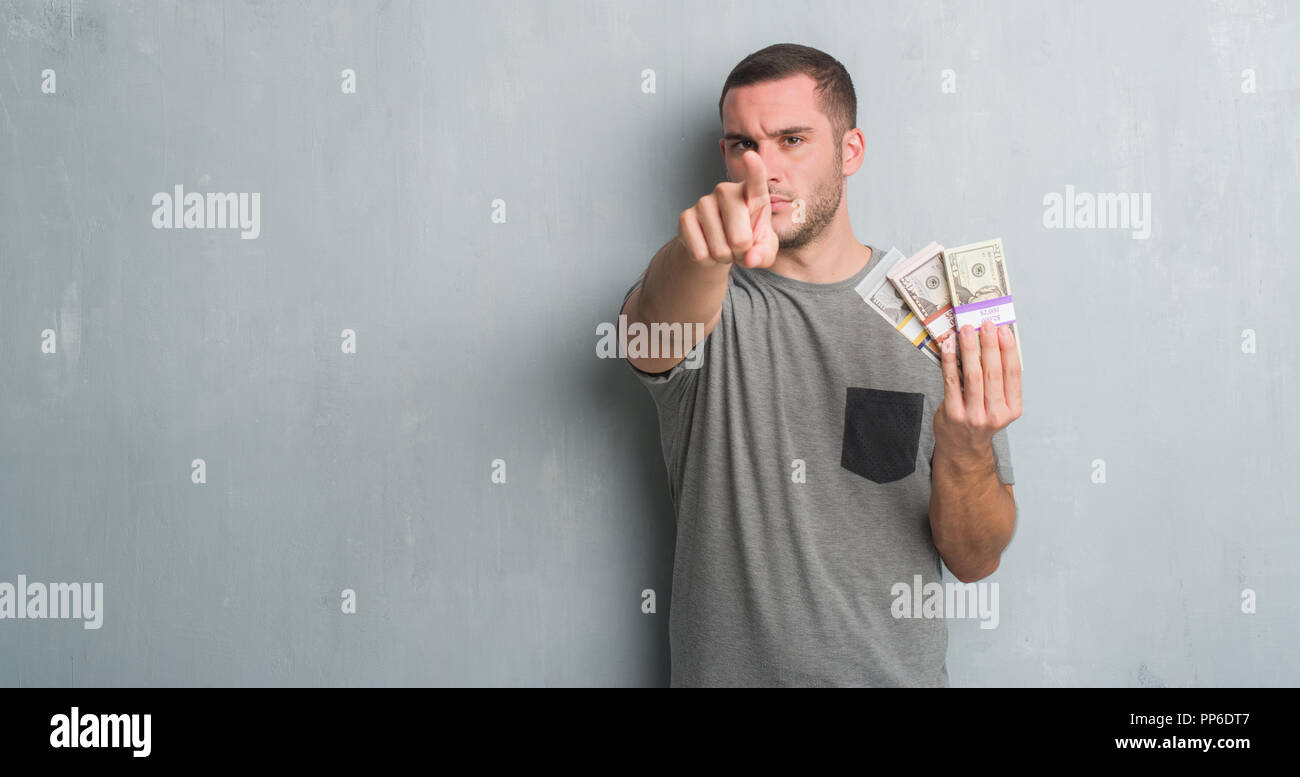 Young caucasian man over grey grunge wall holding a bunch of money pointing with finger to the camera and to you, hand sign, positive and confident ge Stock Photo
