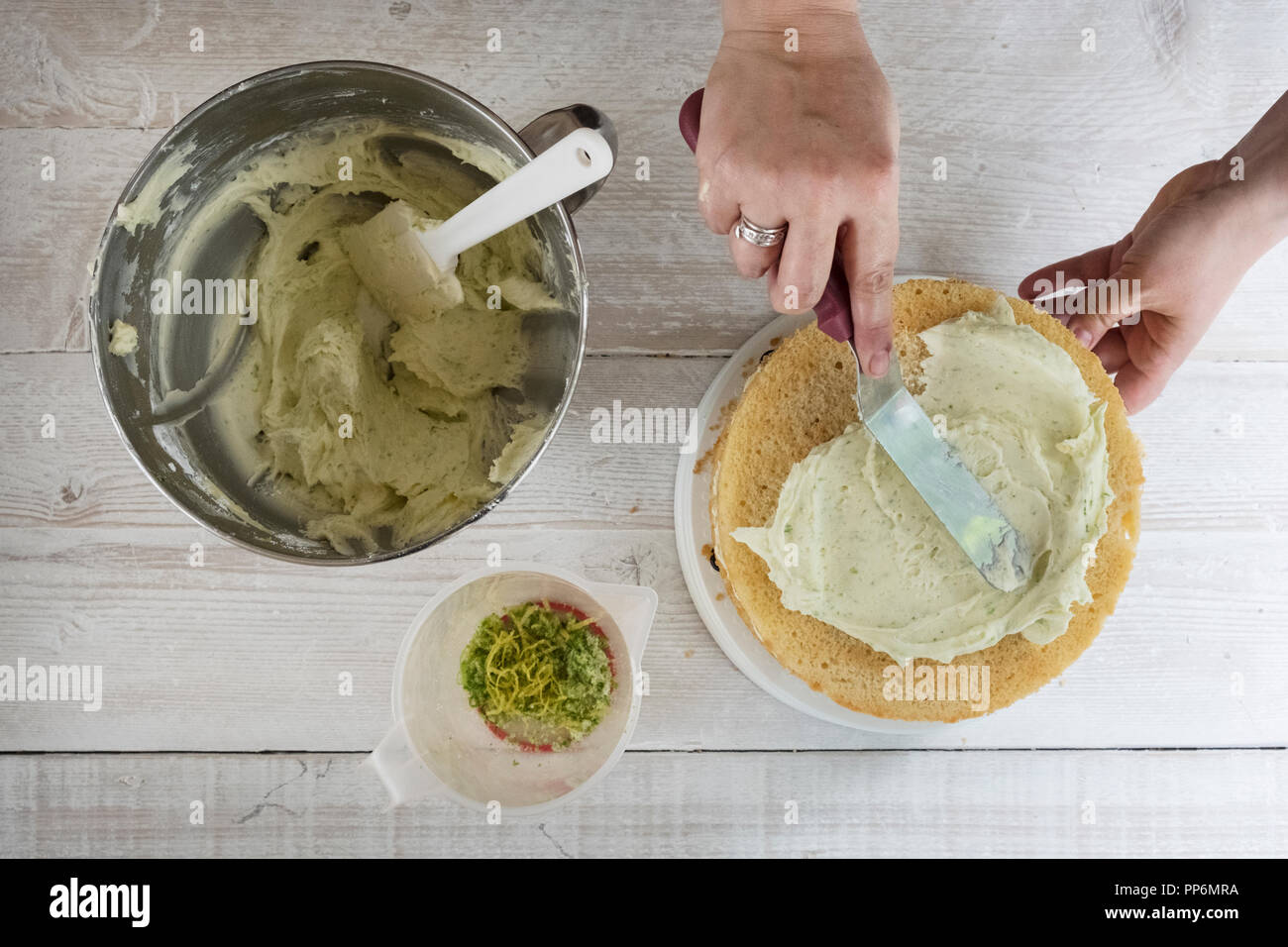 A fresh gin and tonic flavoured baked cake being iced, a hand with a palette knife smoothing icing, and a bowl of fresh icing and lemon zest. Stock Photo