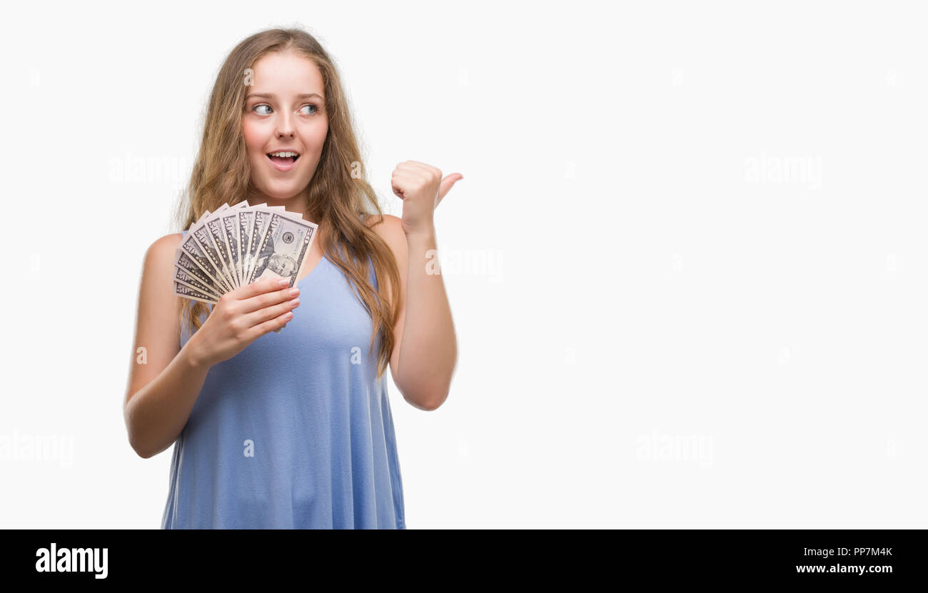 Young blonde woman holding dollars pointing and showing with thumb up to the side with happy face smiling Stock Photo