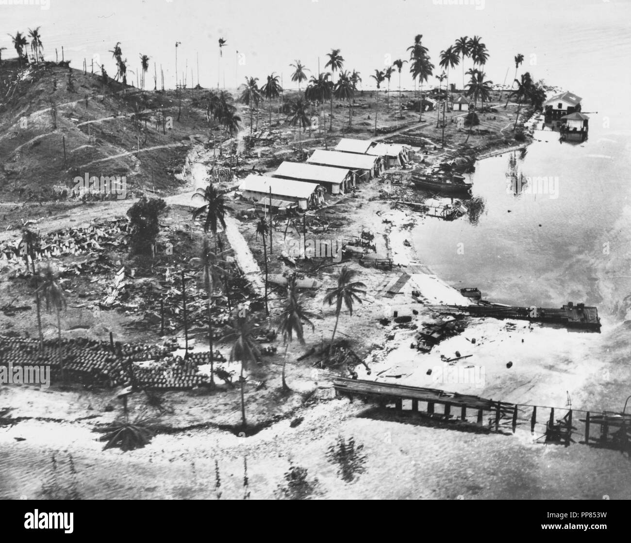 Guadalcanal-Tulagi Landings, 7-9 August 1942.  Wrecked facilities and aircraft at the Japanese seaplane base on Tanambogo Island, east of Tulagi. Photo is dated 8 August 1942 and was probably taken shortly before U.S. Marines captured the island. This view looks about west, with a burned-out pier in the foreground, fuel drums piled to the left and the wreckage of a seaplane among the trees in the center. The buildings are probably left over from the island's days as a Royal Australian Air Force facility. Stock Photo