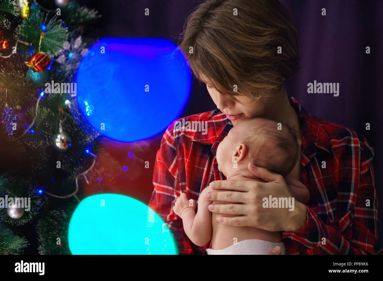 Portrait of young mother and newborn baby. In background Christmas tree and glare from garland. Happiness of motherhood. Stock Photo