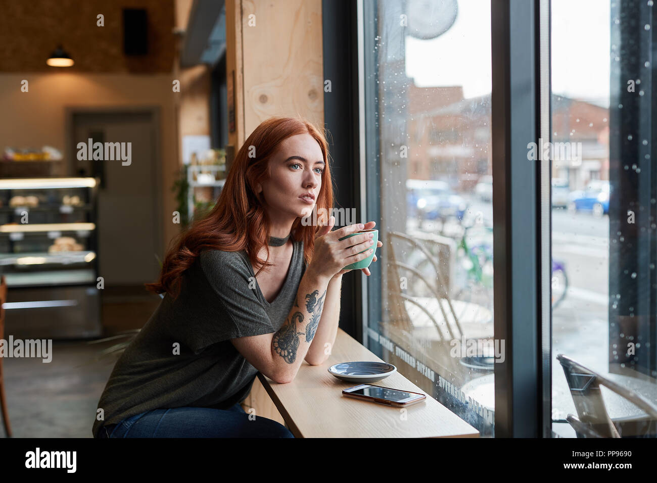 Red Head girl wIth arm Tattoo, sitting alone in a cafe looking through the window, Liverpool, UK Stock Photo