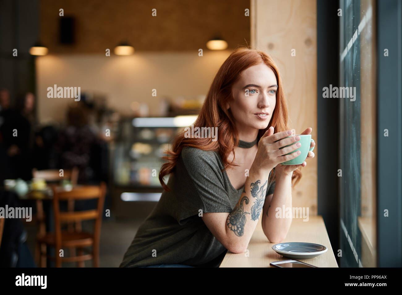 Red Head girl wIth arm Tattoo, sitting alone in a cafe looking through the window, Liverpool, UK Stock Photo