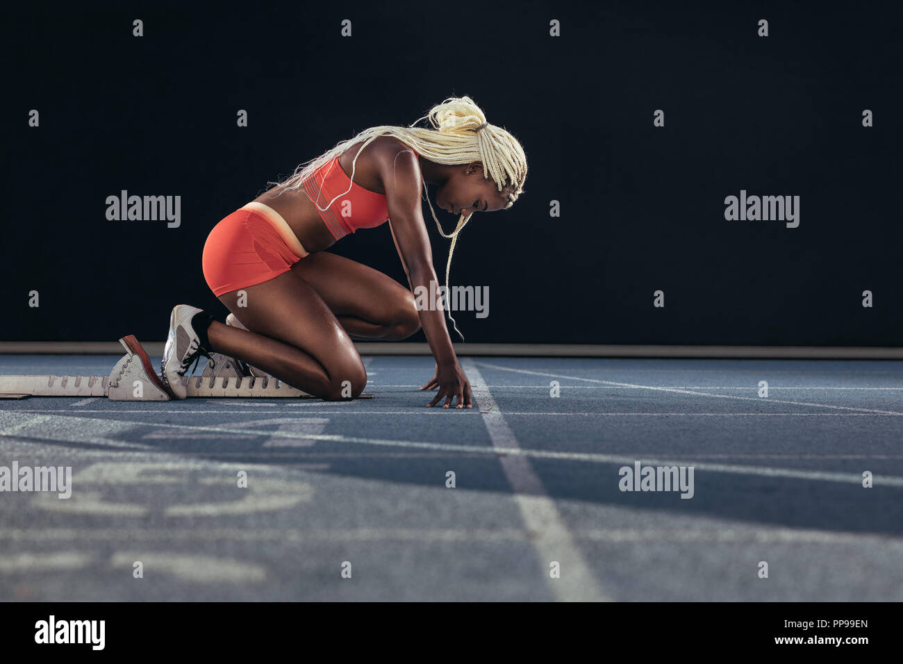 Female athlete taking position on her marks to start off the run. Side view of female runner sitting at the start line on running track on a black bac Stock Photo