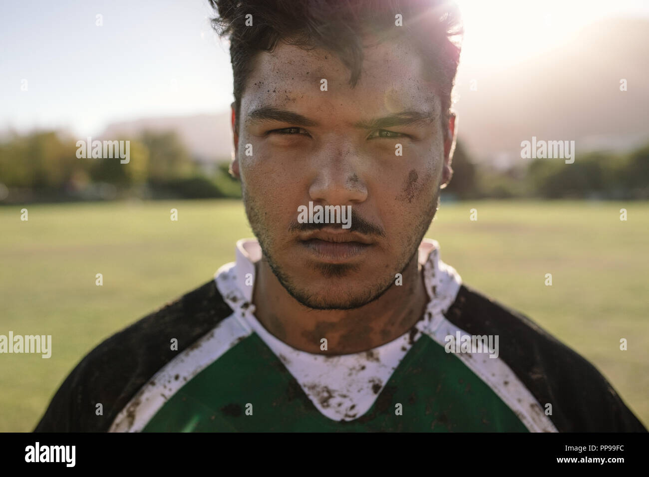 Close up portrait of rugby player with muddy face and uniform on field. Young sportsman staring at camera on ground. Stock Photo