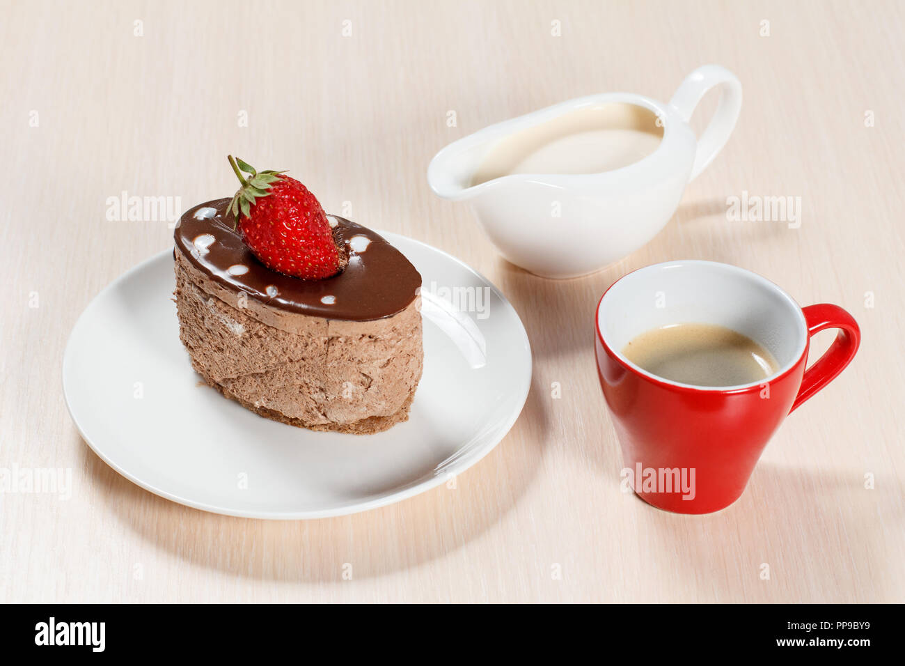 Chocolate cake with strawberry on the white plate, a cup of coffee and a gravy boat with milk on a wooden table Stock Photo