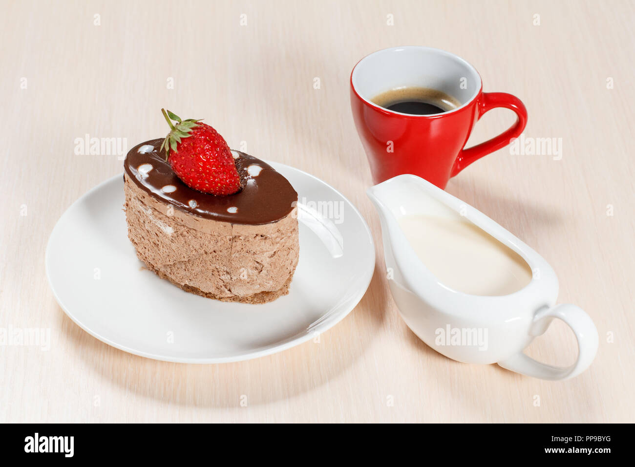 Chocolate cake with strawberry on the white plate, a cup of coffee and a gravy boat with cream on a wooden table Stock Photo