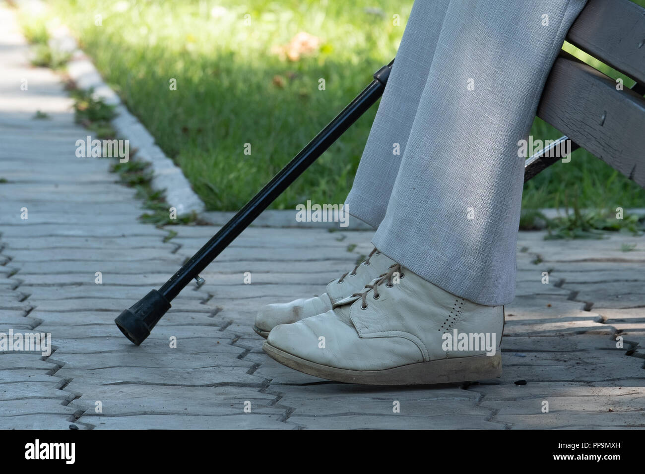 On the image of a foot, a crutch and a bench in the park. On the image of a leg in old pants, shod in old shoes, a crutch and a bench in the park. Stock Photo