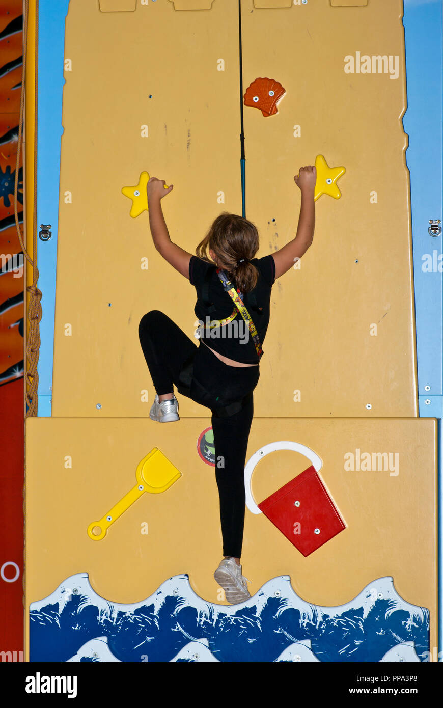 Young Girl Striving To Climb a Climbing Wall Secured By A Safety Harness Stock Photo