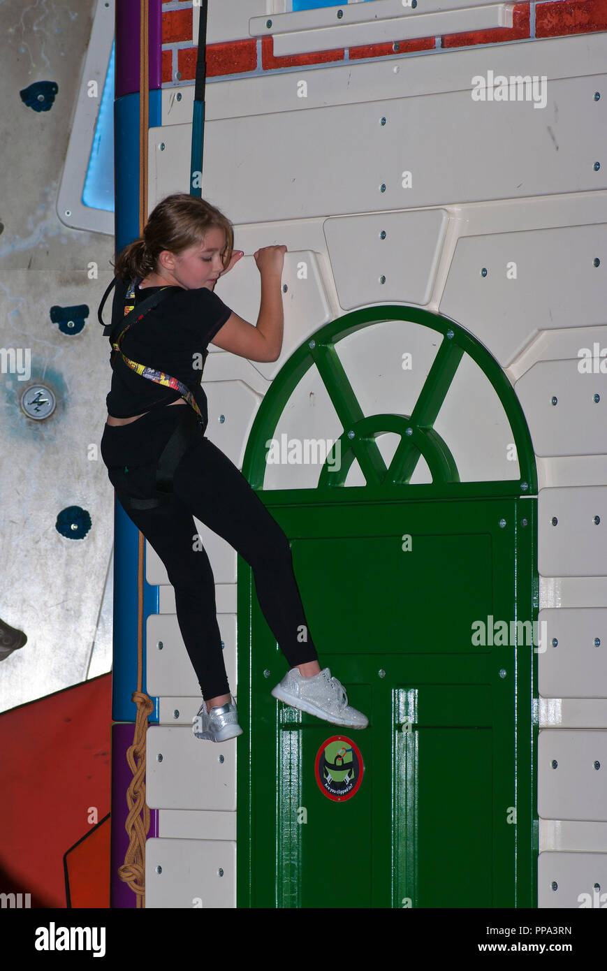 Young Girl Striving To Climb a Climbing Wall Secured By A Safety Harness Stock Photo