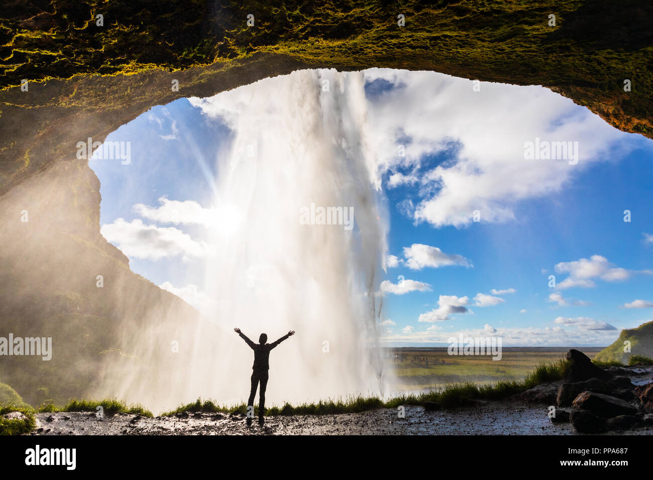 Happy female traveler feeling the power of Seljalandsfoss waterfall in the South of Iceland, person standing behind the stream, famous Icelandic landm Stock Photo