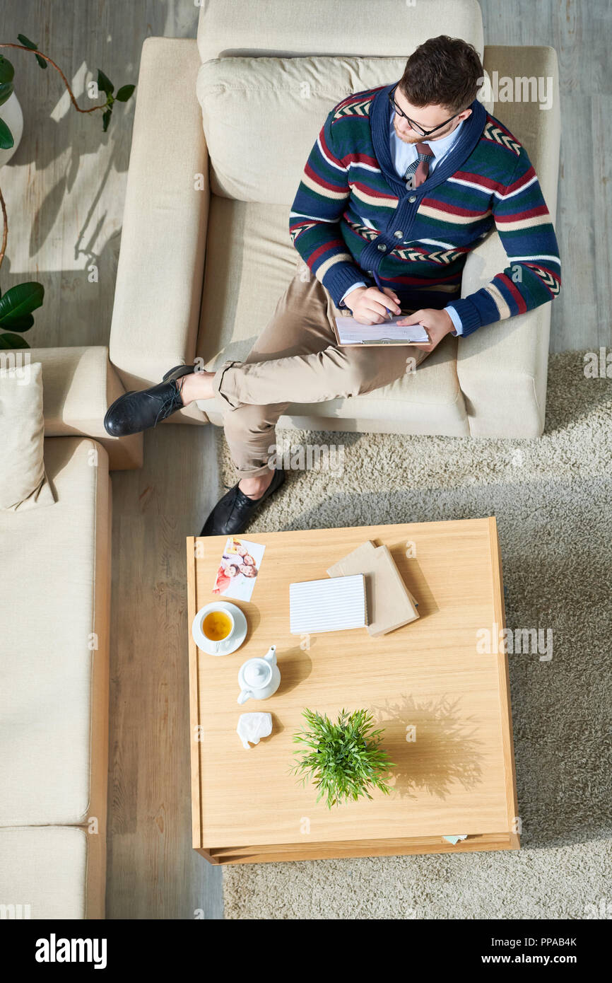Above view portrait of pensive male psychologist writing notes on clipboard while sitting in comfortable armchair in sunlit office, copy space Stock Photo