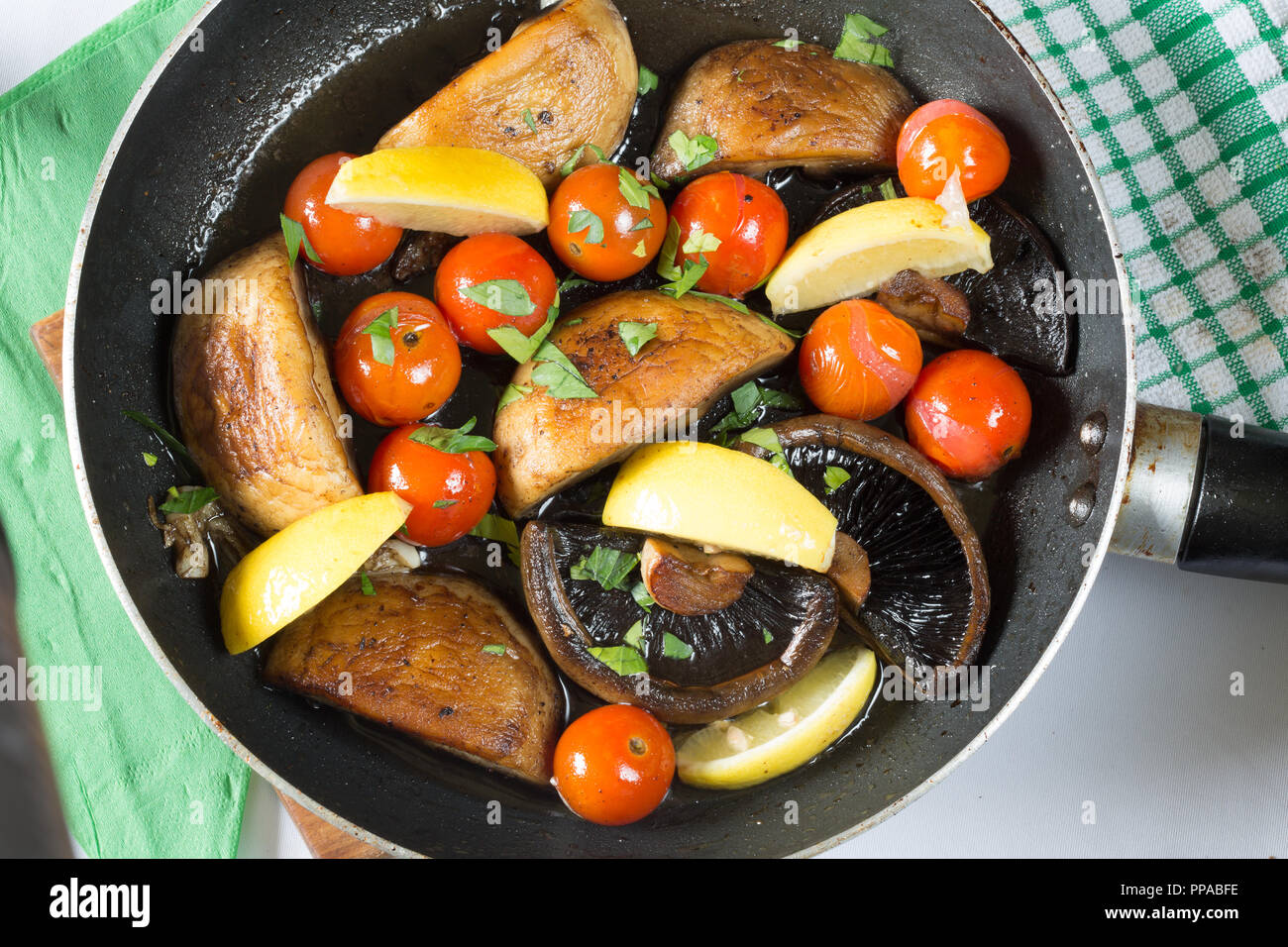 Mushrooms and Cherry Tomatoes being stir fried in a shallow frying pan, garnished with fresh chopped Parsley and Lemon wedges Stock Photo