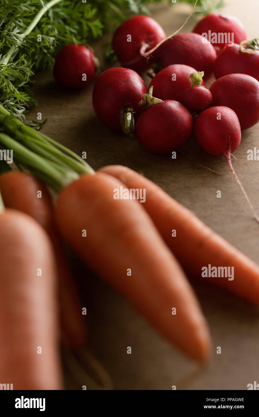 Close-up of a small bunch of radishes, surrounded by fresh carrots and a head of lettuce on a light brown board, under morning light. Stock Photo