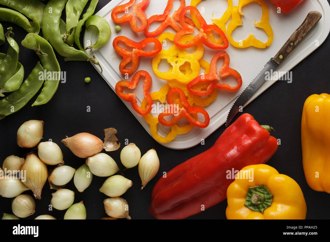 Ripe bell peppers slices on a white chopping board, surrounded by other vegetables, like onions and snow peas on a black table. Stock Photo