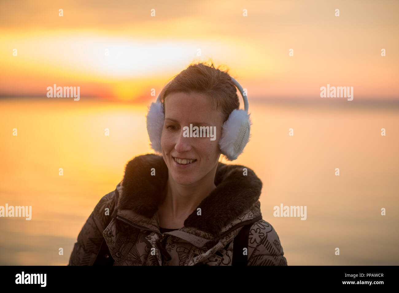 Smiling woman wearing warm fluffy ear muffs at sunset standing against a backdrop of a calm ocean and fiery vibrant orange sky as she enjoys a winter  Stock Photo