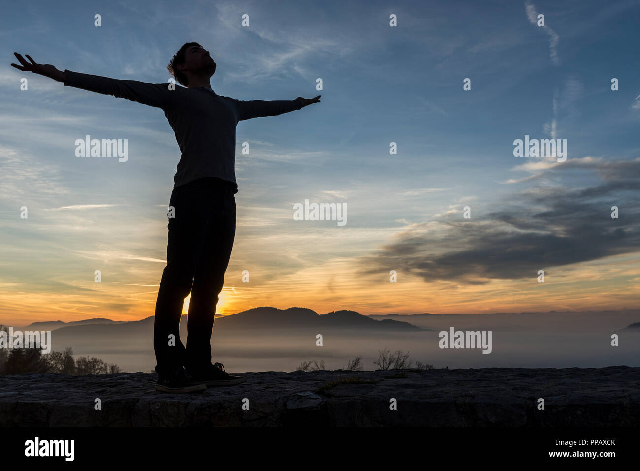 Man in casual clothes with his arms spread widely standing outdoors silhouetted against a sunrise with the fiery sun peeping over mountains. Stock Photo