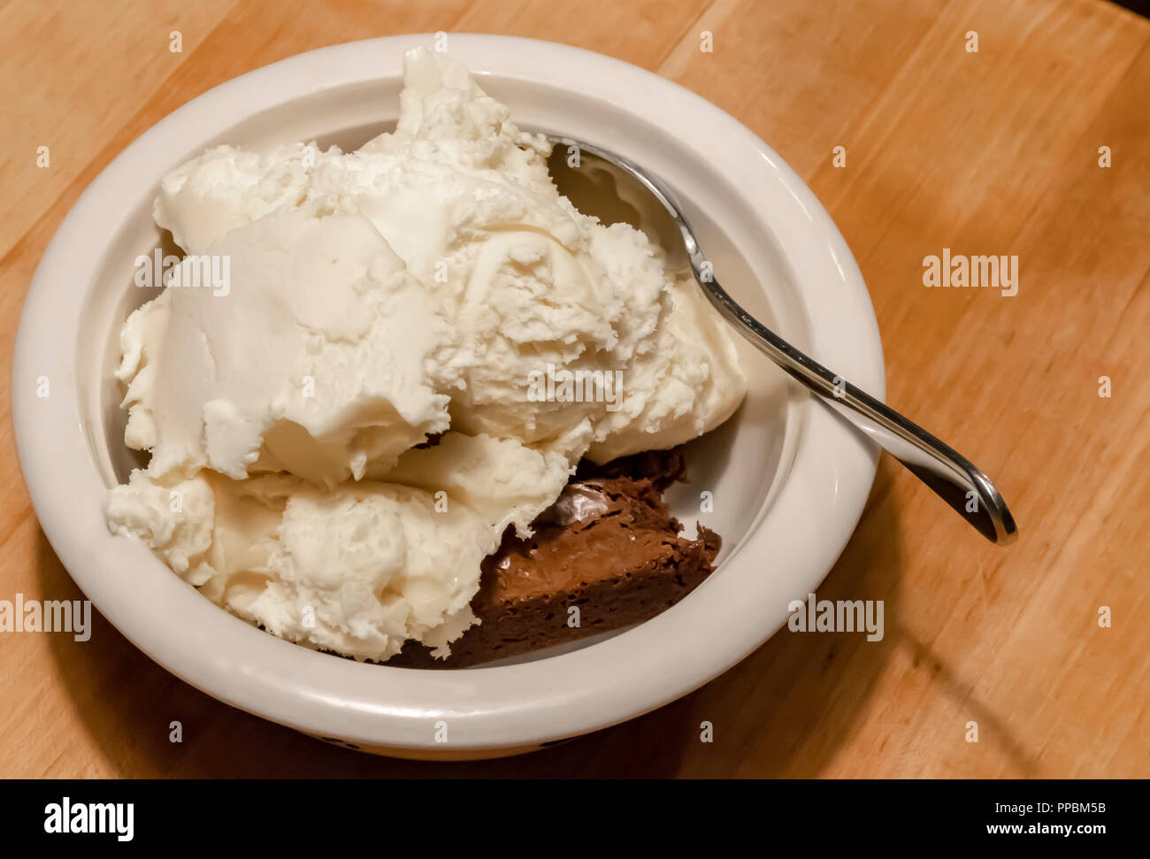 Ice cream and a spoon almost cover a brownie in a bowl on a wooden countertop. Stock Photo