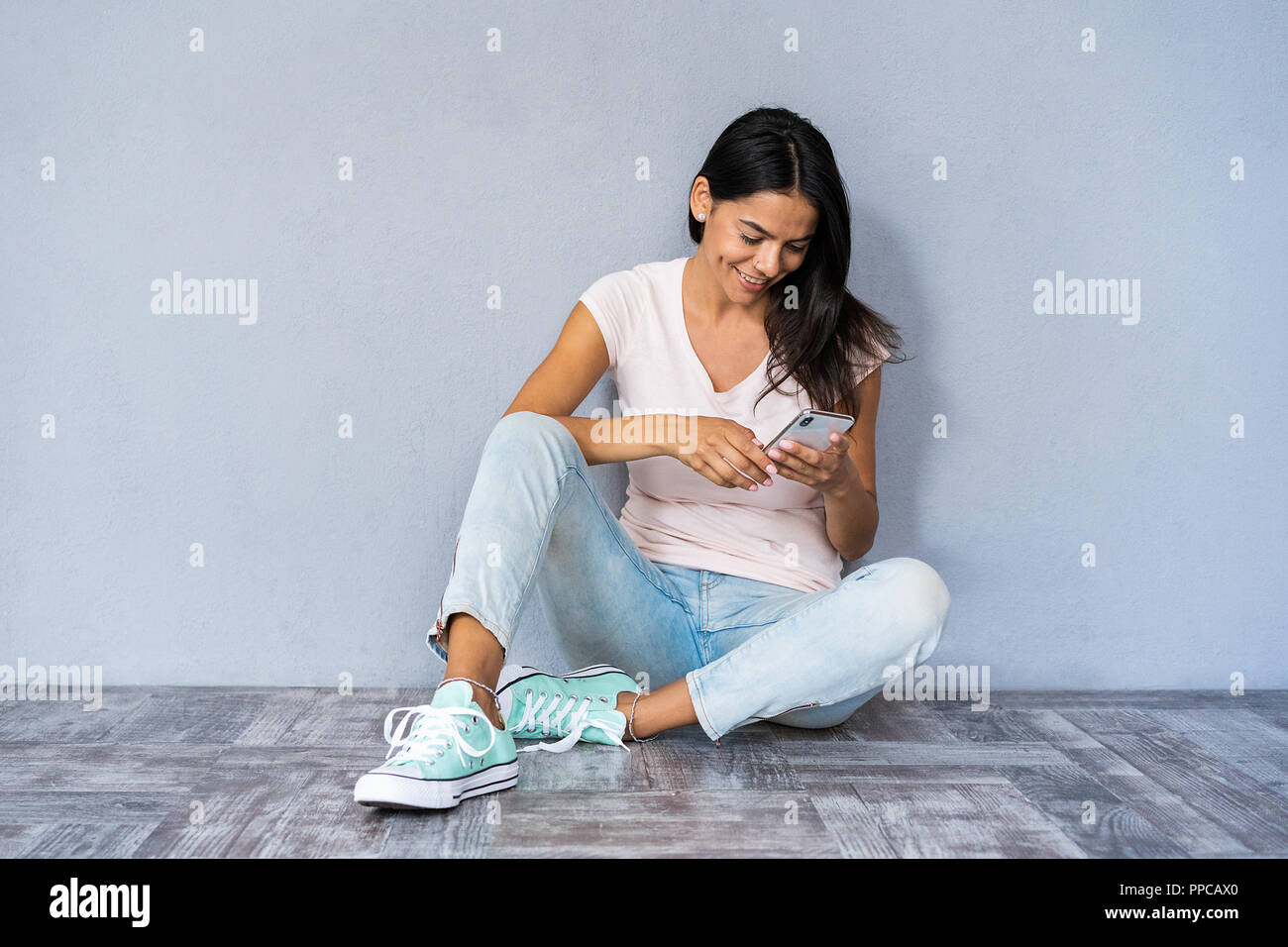 Happy brunette woman sitting on the floor and writing message on smartphone over gray background. Stock Photo